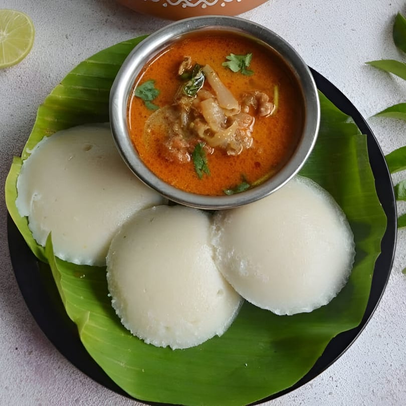 Three white idlis and a bowl of orange curry with meat and cilantro on a banana leaf.