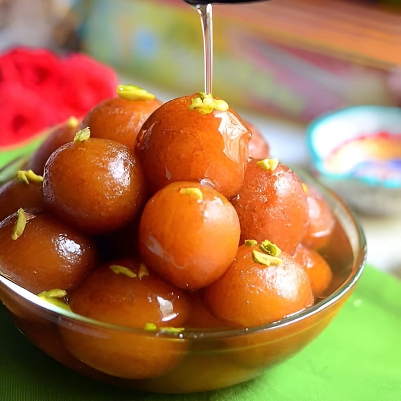 Gulab Jamun, an Indian dessert, in a glass bowl, with syrup being poured over them.