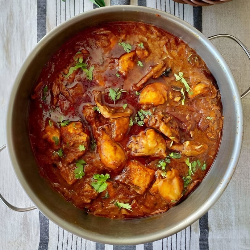A large metal bowl filled with rich, dark red Indian chicken curry, garnished with fresh cilantro.